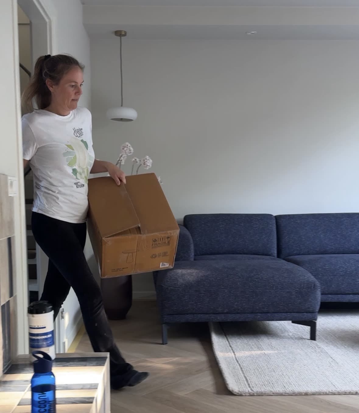 Sharon draagt een doos naar binnen tijdens het inrichten, met de nieuwe bank en lamp al op hun plek